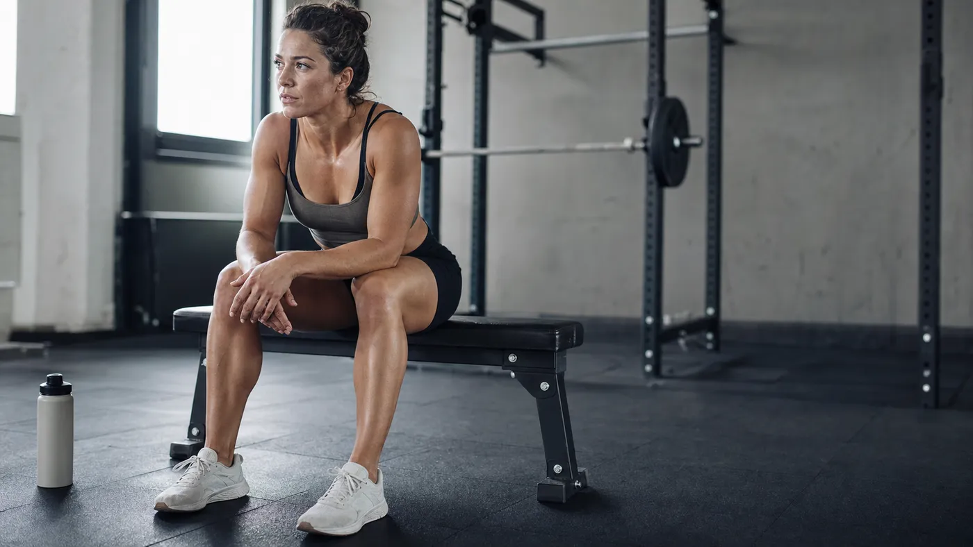 A muscular woman resting calmly on a bench with a water bottle after a hard gym set